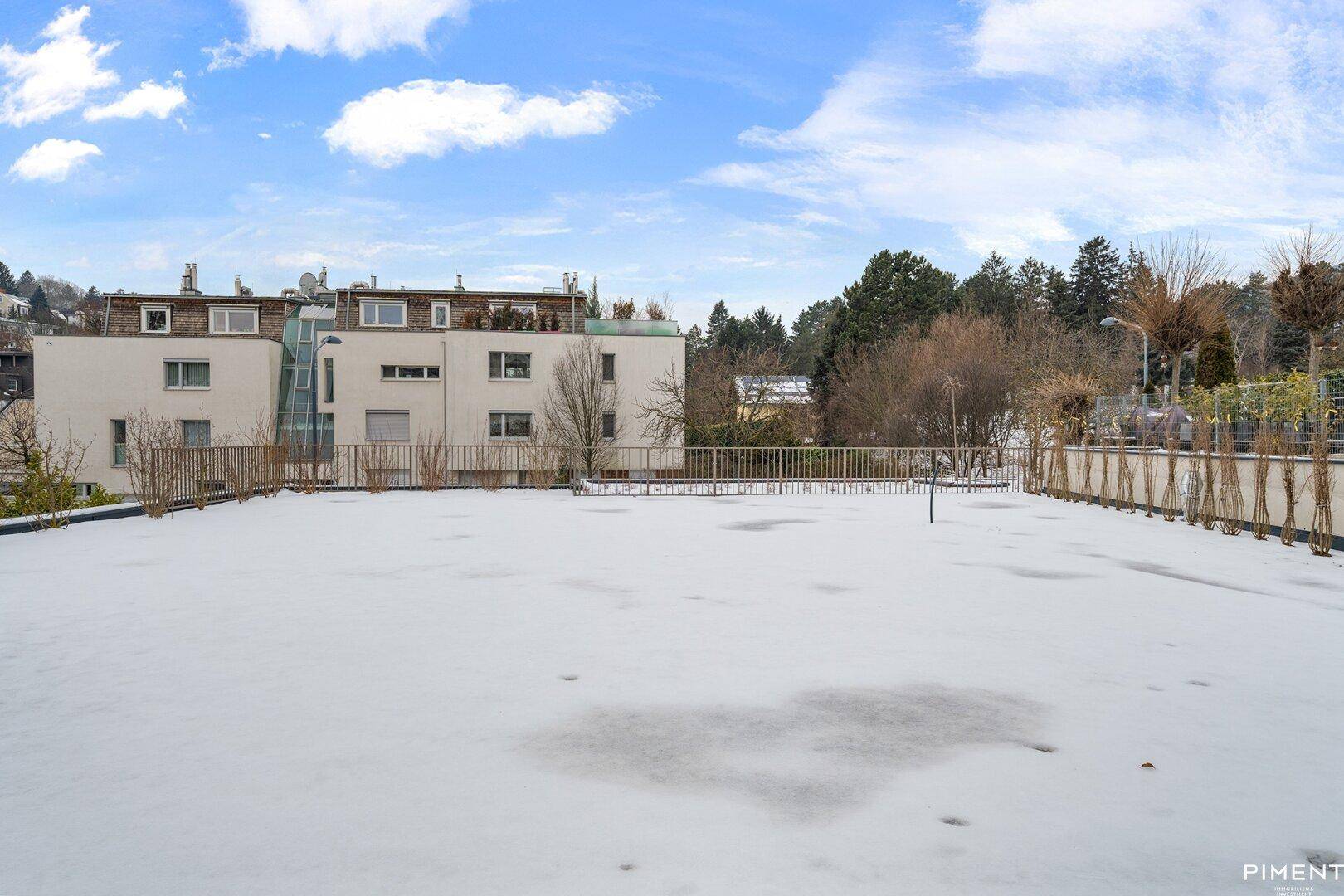 Weitläufige, verschneite Gartenfläche mit Blick auf umliegende Wohngebäude unter blauem Himmel.