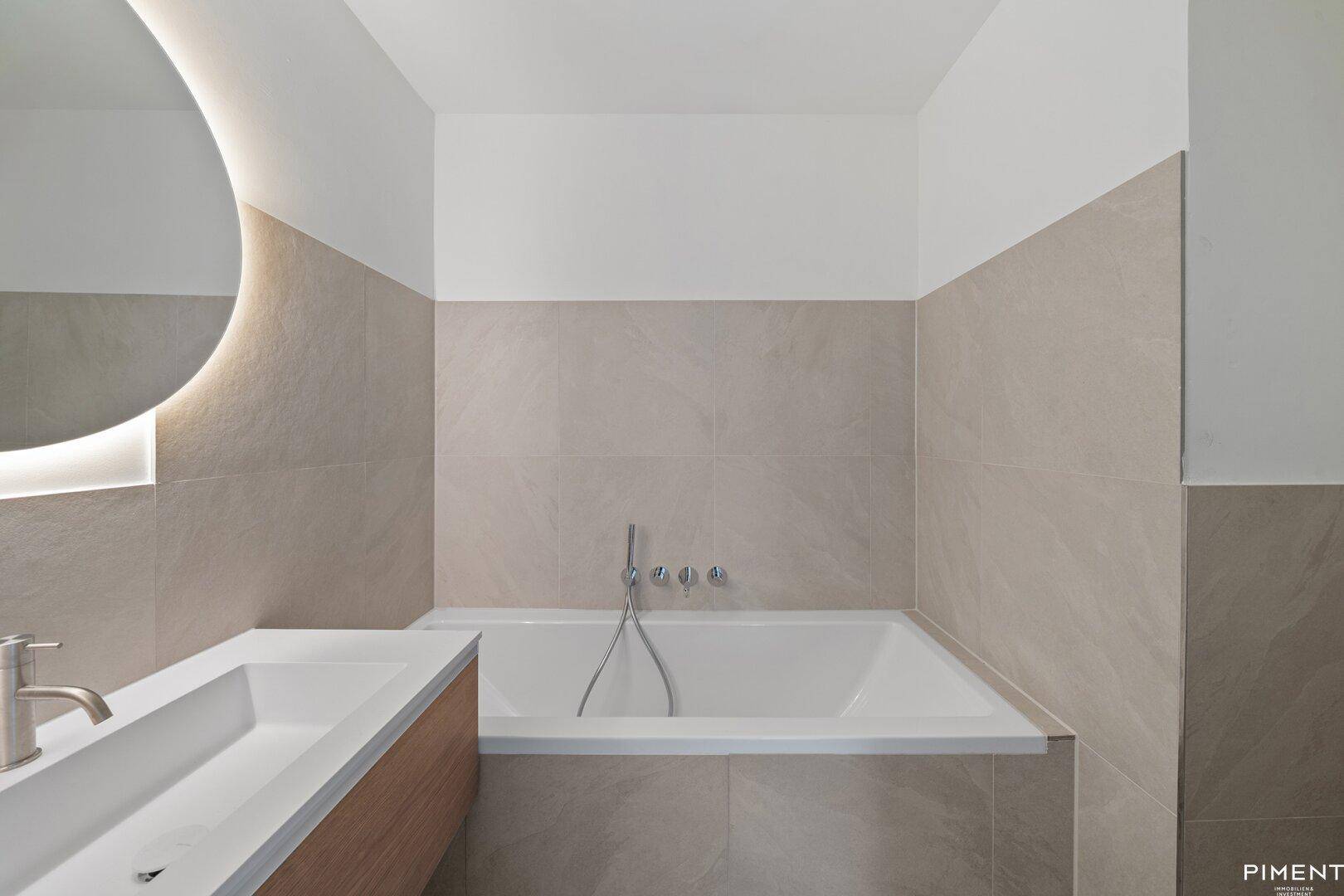 Modernes Badezimmer mit integrierter Badewanne, eleganten Fliesen und beleuchtetem Spiegel.