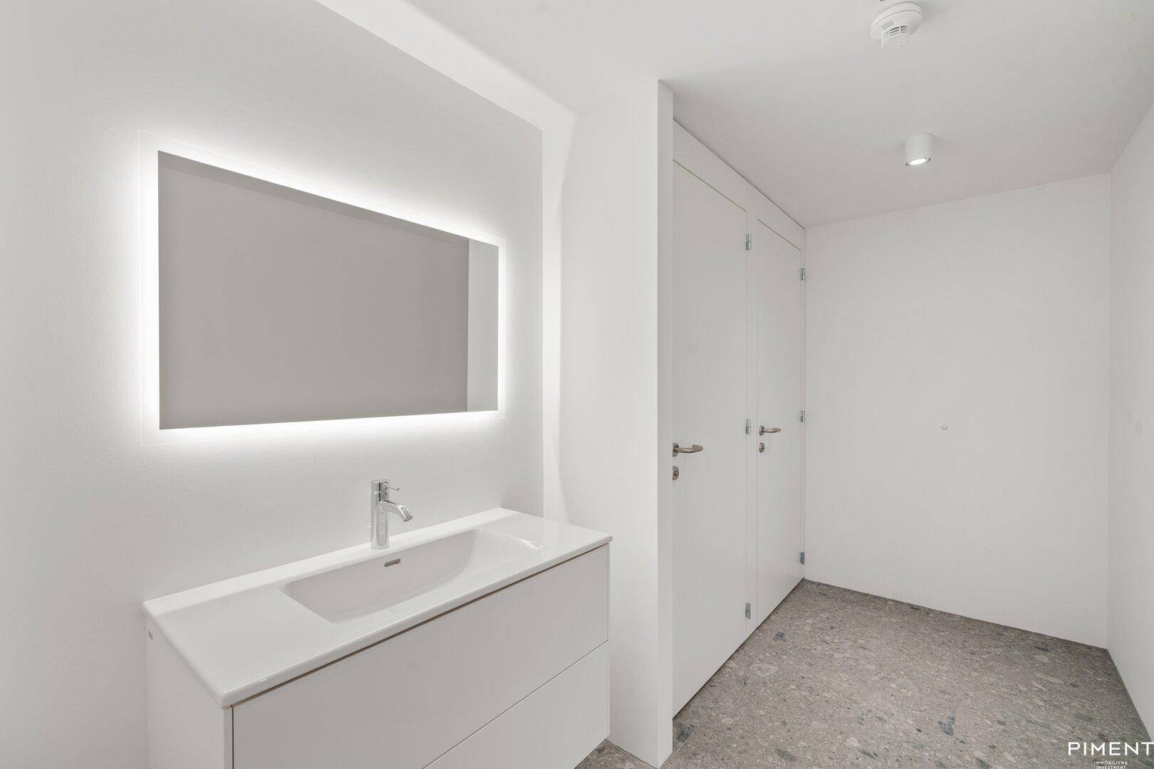 Modernes Badezimmer mit beleuchtetem Spiegel, weißem Waschtisch und minimalistischem Design.
