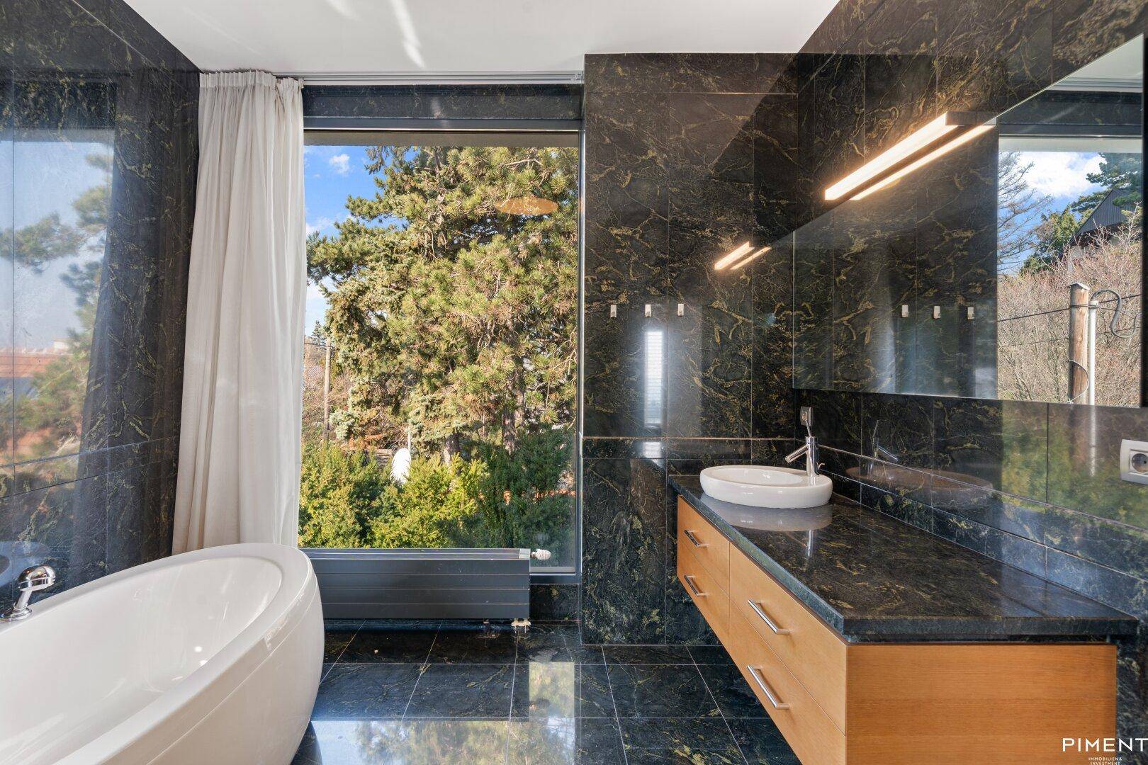Luxuriöses Badezimmer mit dunklen Marmorfliesen, freistehender Badewanne und großem Fenster mit Blick ins Grüne.