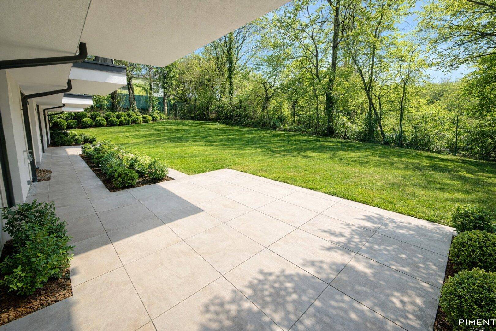 Großzügige, geflieste Terrasse mit direktem Zugang zum gepflegten Rasen und umgebenden Bäumen.