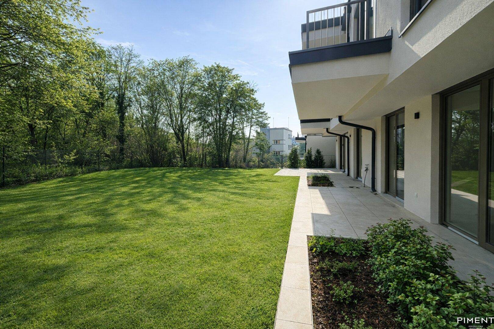Weitläufige, geflieste Terrasse neben einem großen Rasen, umgeben von üppiger Vegetation.
