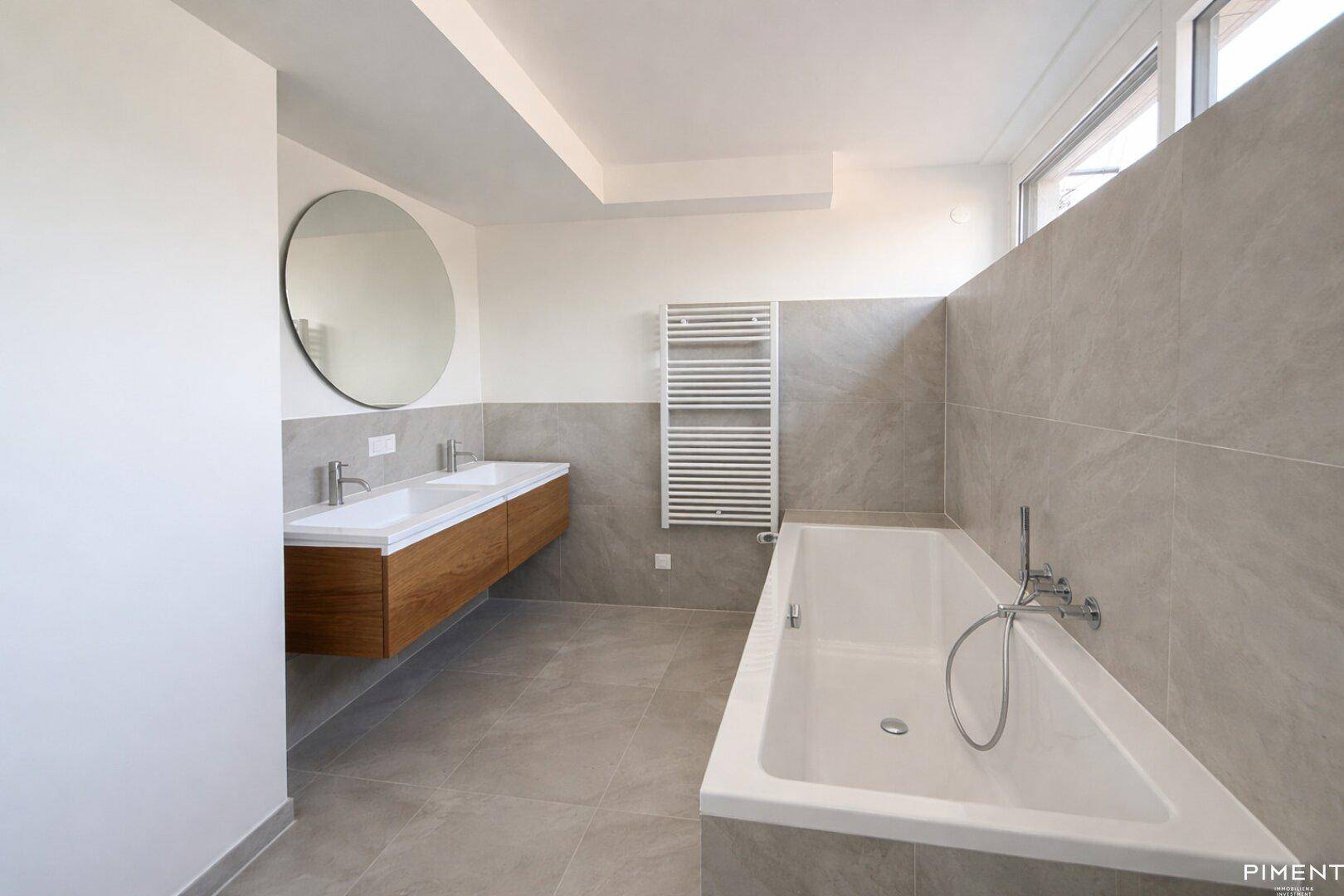 Modernes Badezimmer mit Doppelwaschbecken, Badewanne, Handtuchheizkörper und großen Fliesen.