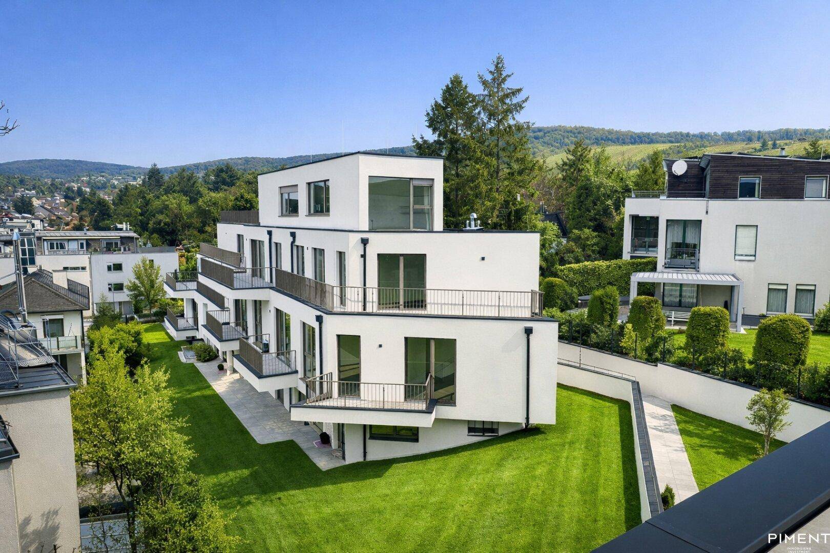 Luftaufnahme eines modernen Wohnkomplexes mit Flachdach und Terrassen, eingebettet in grüne Hügellandschaft.