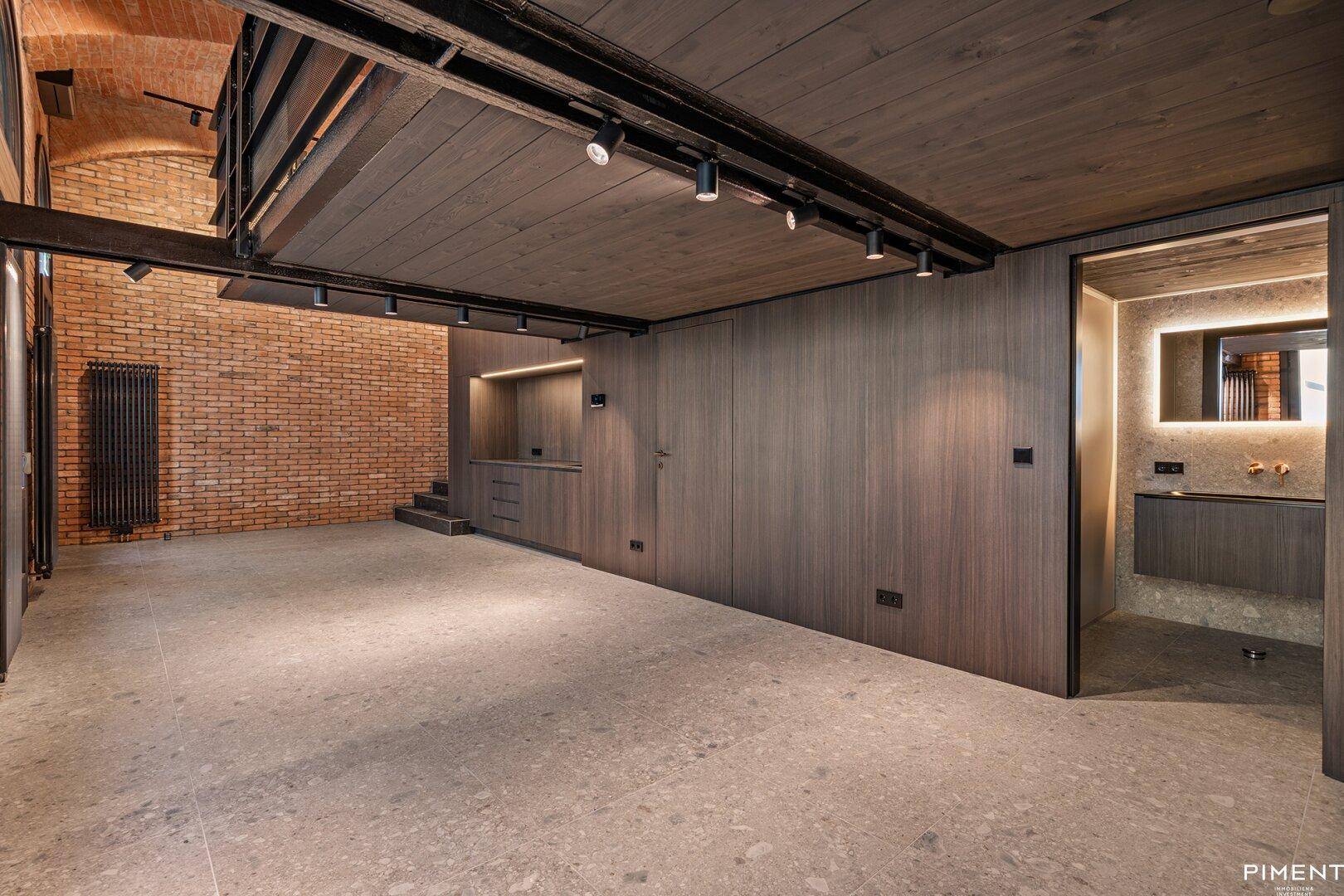 Geräumiger Loft-Bereich mit Ziegelwänden, Gewölbedecke und Zugang zu einem modernen Badezimmer.