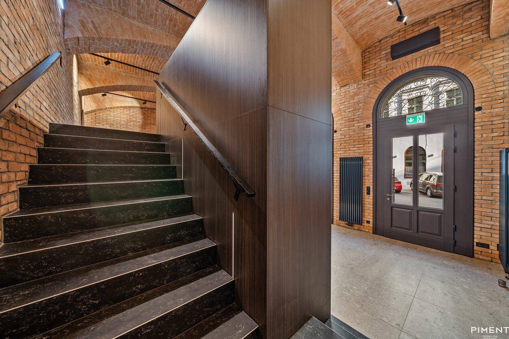 Elegantes Treppenhaus mit dunklen Stufen, freiliegenden Ziegelwänden und einer beeindruckenden Gewölbedecke.