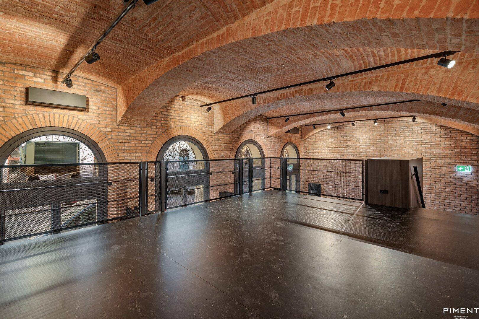 Geräumige Galerieebene mit Ziegelwänden, Gewölbedecke und mehreren Rundbogenfenstern mit Blick nach draußen.