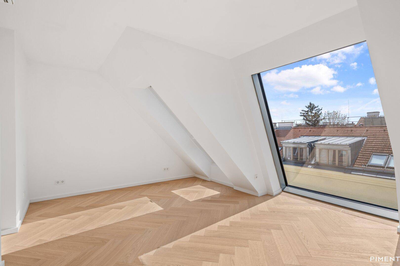 Helles Zimmer im Dachgeschoss mit großem Fenster und Blick auf die umliegenden Dächer.