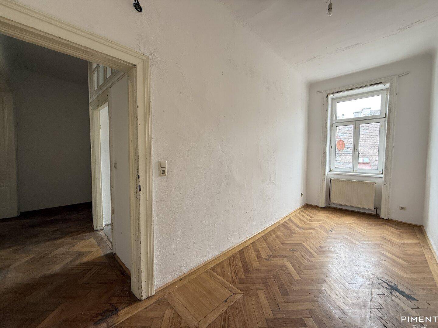 Helles Zimmer mit Fischgrätparkett und Zugang zu einem weiteren Raum, renovierungsbedürftig.