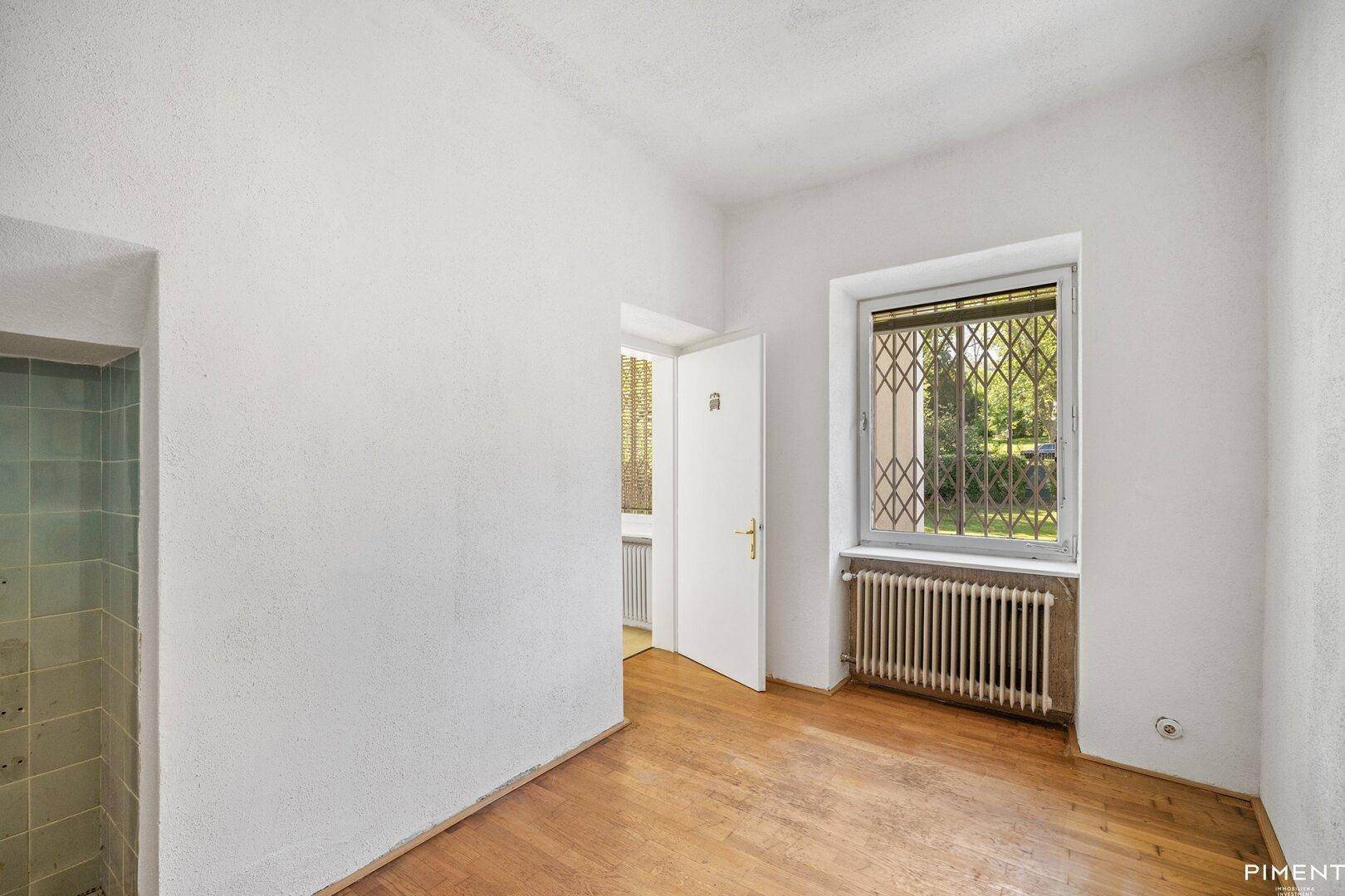 Heller Raum mit Parkettboden und einem Fenster mit Sicherheitsgitter.