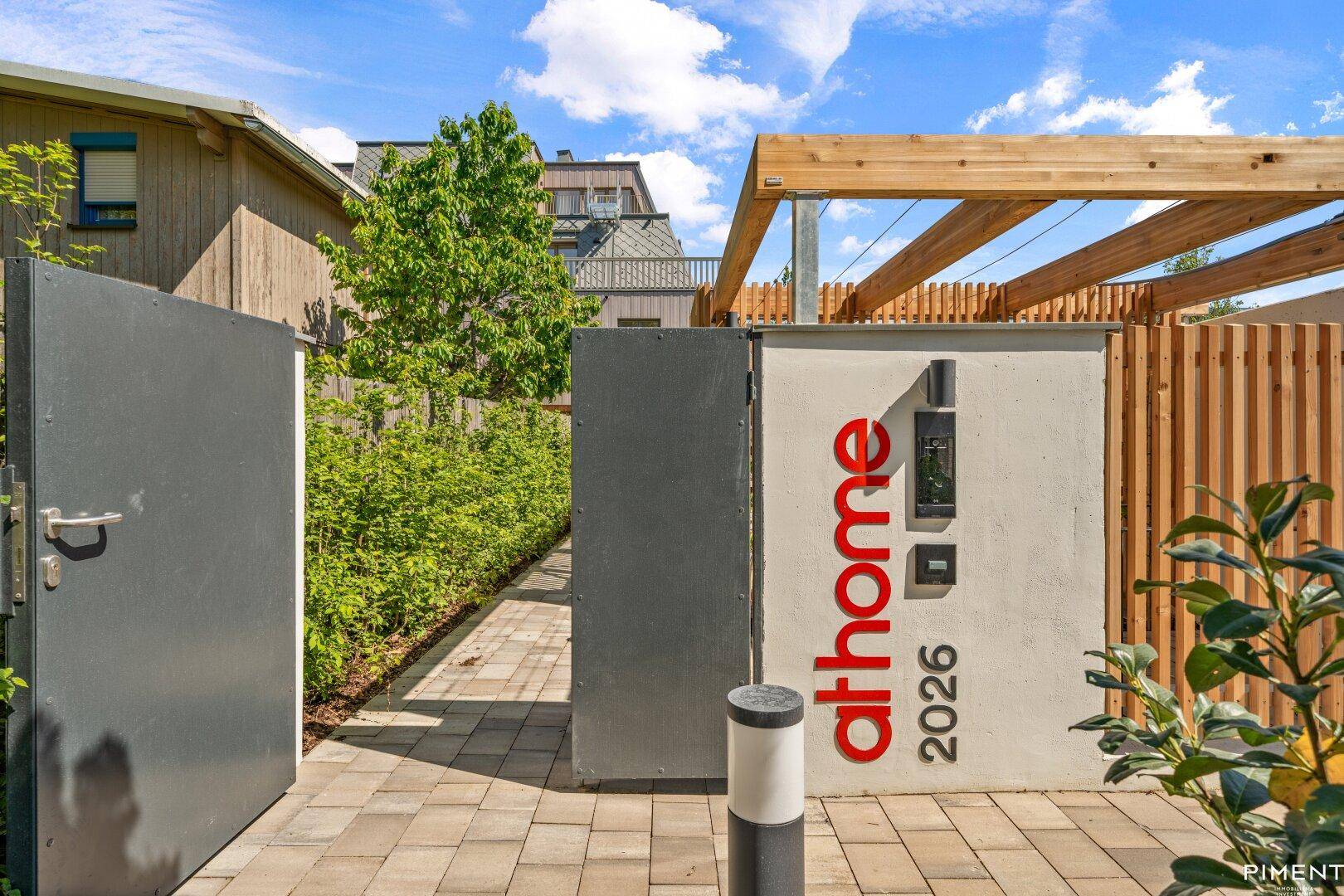 Eingangsbereich mit einem modernen Tor, einer Pergola und einem Hinweisschild zur Wohnanlage.