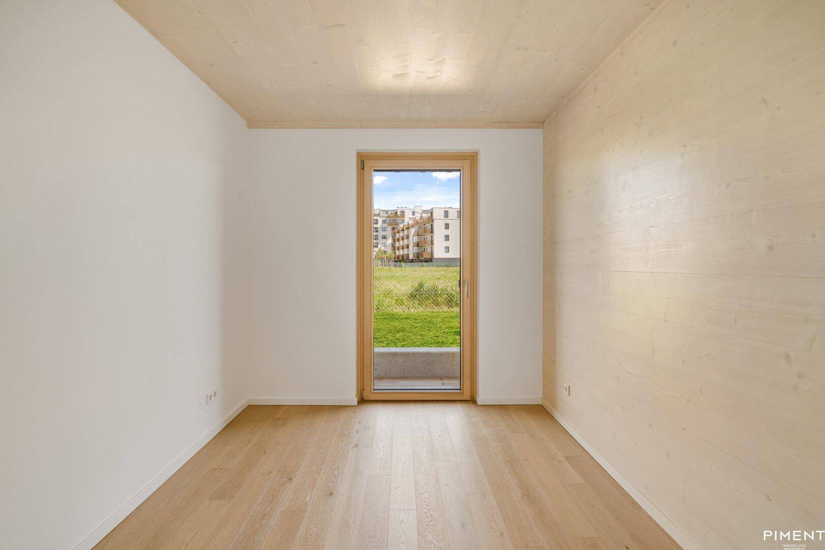 Unmöbliertes Zimmer mit Holzboden und einer Glastür, die einen Blick auf die umliegende Grünfläche bietet.