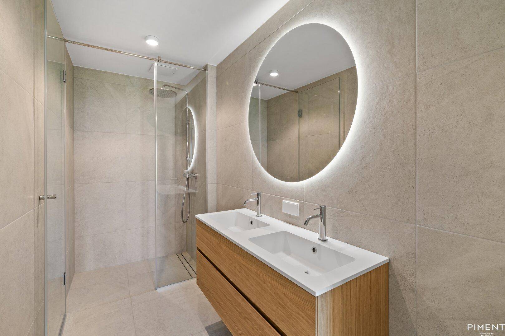 Modernes Badezimmer mit Doppelwaschbecken, beleuchtetem Spiegel und einer geräumigen begehbaren Dusche.