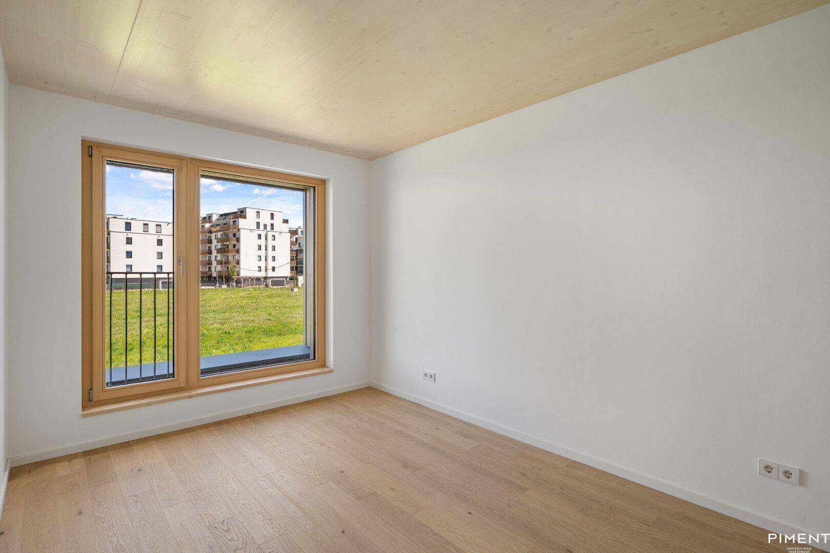 Helles Zimmer mit Holzboden und Fenster mit Blick auf eine Grünfläche und Gebäude.