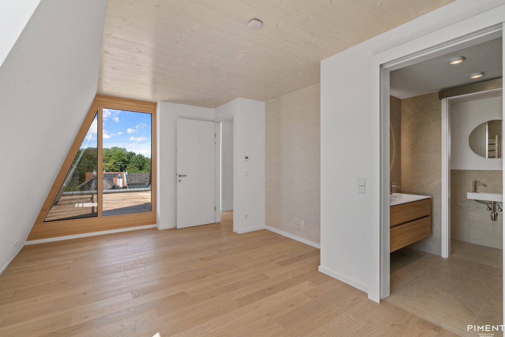 Helles Zimmer mit Holzboden, großem Fenster und angrenzendem modernen Badezimmer.