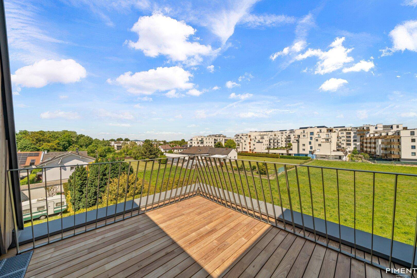 Heller Holzbalkon mit Metallgeländer und weitem Blick über eine Wiese und Gebäude.