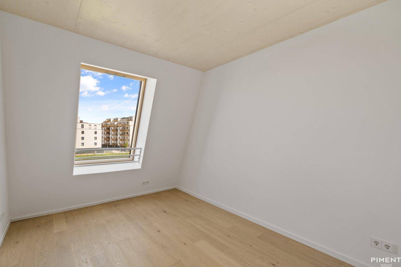 Helles Schlafzimmer mit Holzboden, Dachfenster und Blick auf die umliegenden Gebäude.