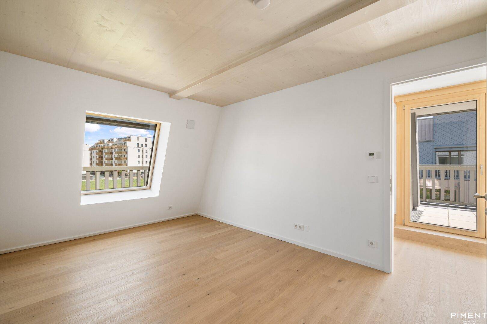 Helles, unfurniertes Zimmer mit Holzboden, schrägem Fenster und Zugang zu einem Balkon.