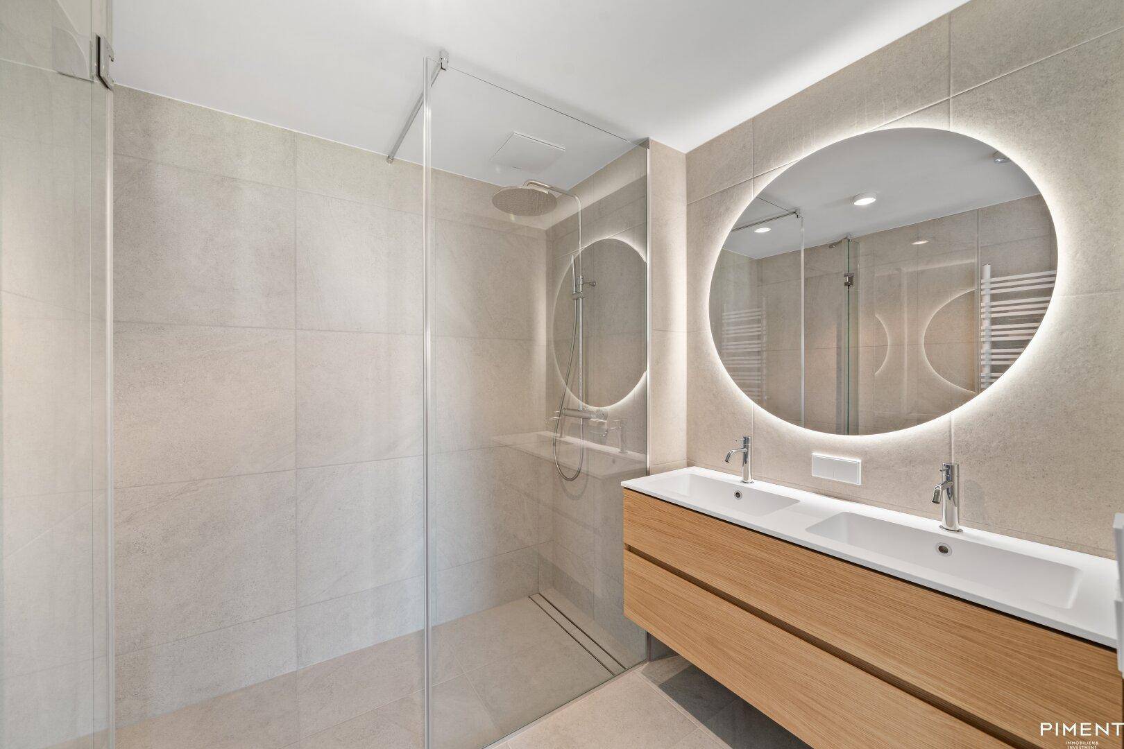 Modernes Badezimmer mit Doppelwaschbecken, beleuchtetem Spiegel und großer begehbarer Dusche.