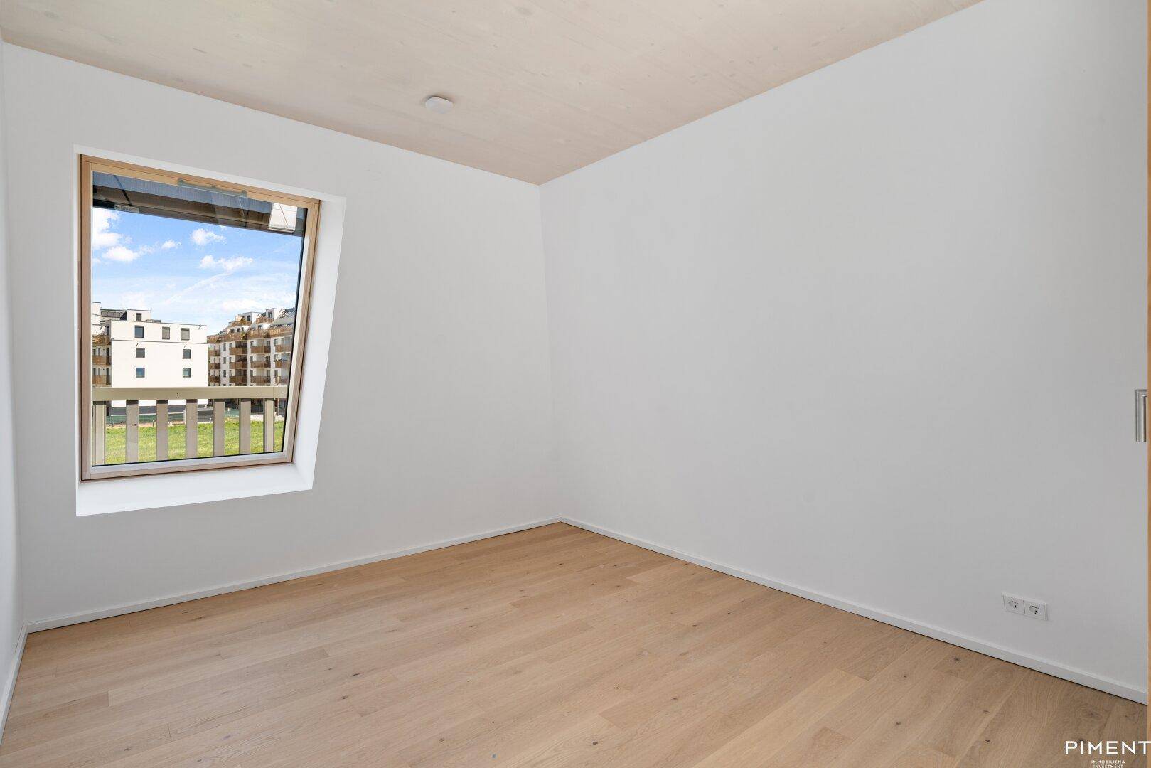 Unmöbliertes Schlafzimmer mit Holzboden und schrägem Fenster, das viel Tageslicht hereinlässt.