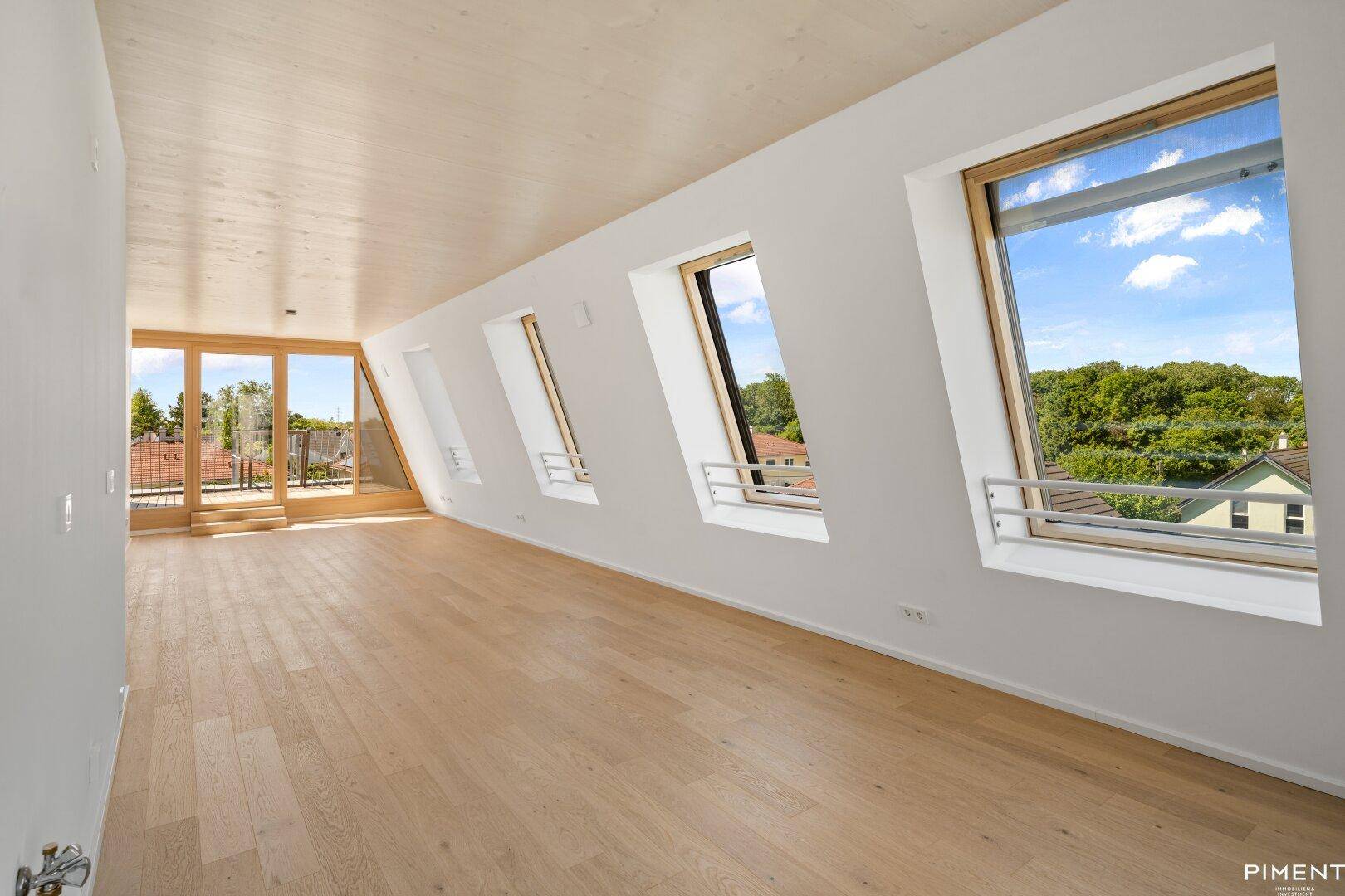 Heller Raum mit Holzboden, Dachschrägenfenstern und Zugang zu einem Balkon mit grüner Aussicht.