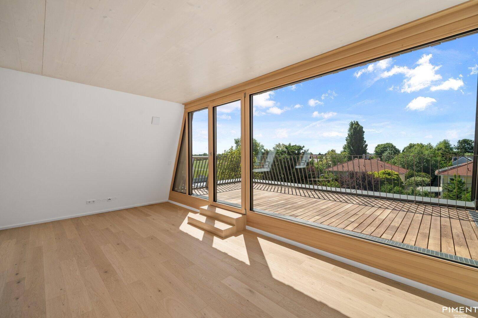 Lichtdurchfluteter Raum mit Holzboden und breiter Fensterfront zur Holzterrasse.