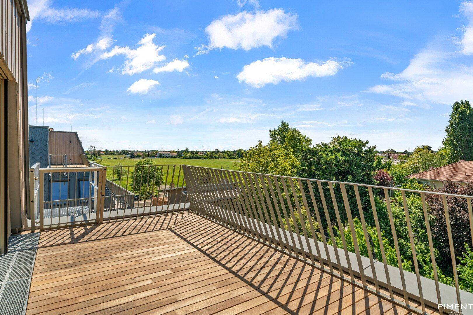 Große Holzterrasse mit Metallgeländer und weitem Blick über grüne Felder und Dächer.