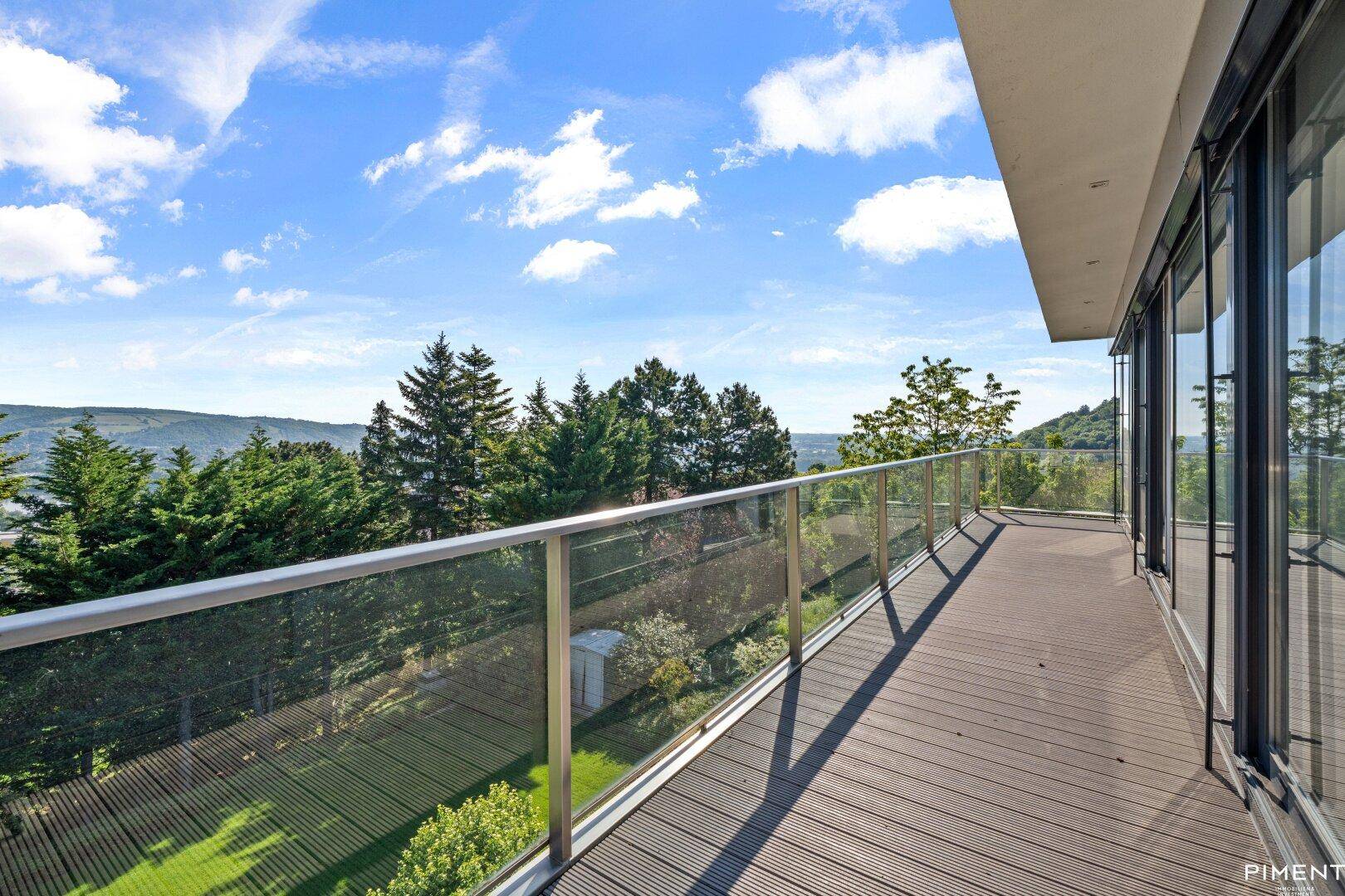 Langer Balkon mit Glasgeländer und Holzboden, bietet einen weiten Blick über die Baumwipfel und die Landschaft.