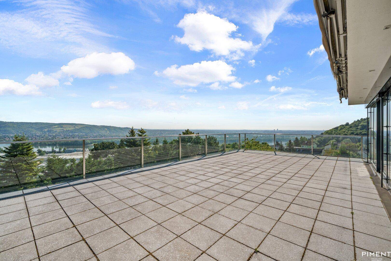 Große Terrasse mit Glasgeländer und weitem Blick über das Tal und den Fluss unter blauem Himmel.