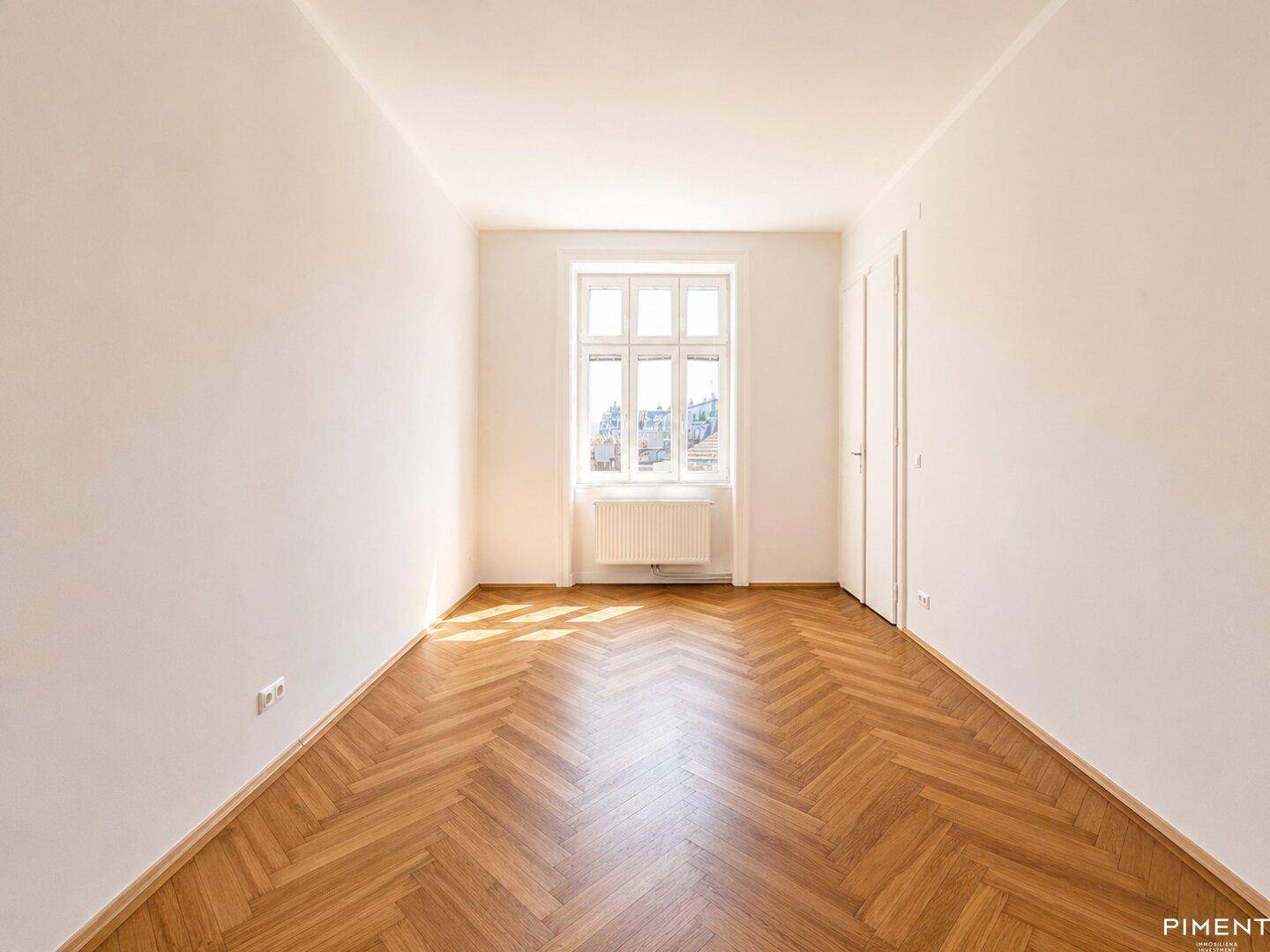 Ruhiges Zimmer mit Fischgrätparkett, einem Fenster und zwei weißen Türen, ideal als Schlafzimmer.