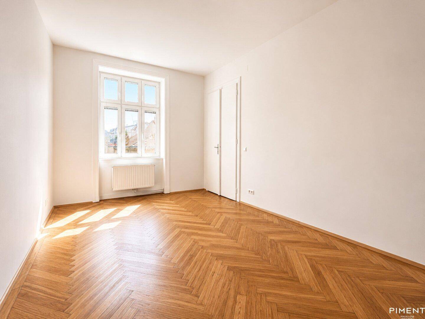 Sonniger Raum mit klassischem Fischgrätparkett und einem großen Fenster, das viel Licht hereinlässt.
