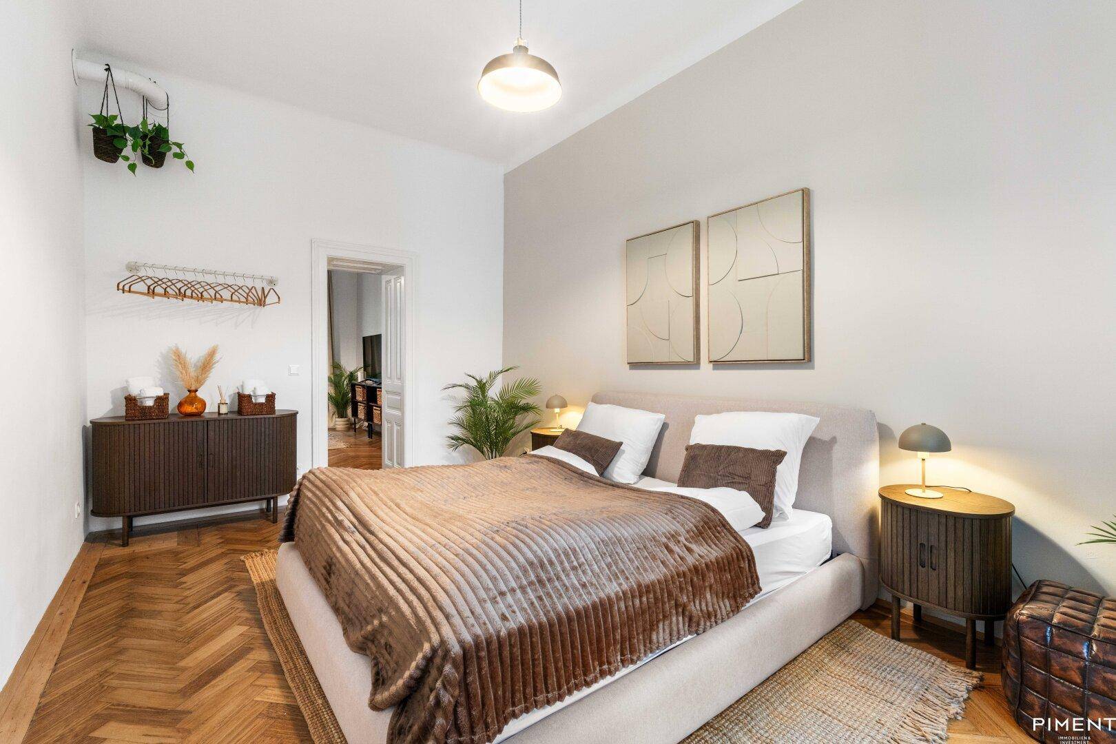 Helles Schlafzimmer mit beigem Bett, Holzboden und Blick in den Wohnbereich.