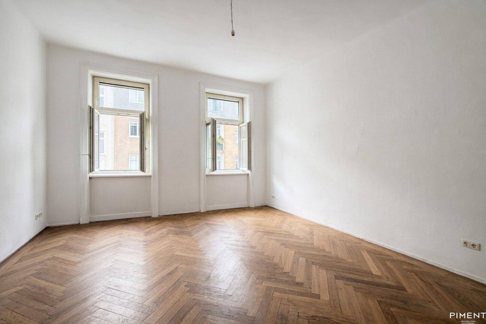 Geräumiges, leeres Zimmer mit Fischgrätparkett und zwei großen Fenstern.