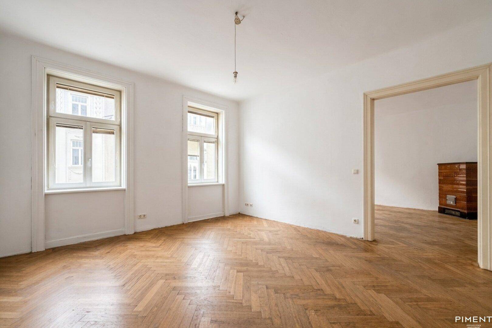 Leeres Zimmer mit Fischgrätparkett, hohen Decken und Durchgang zu einem Nebenraum.