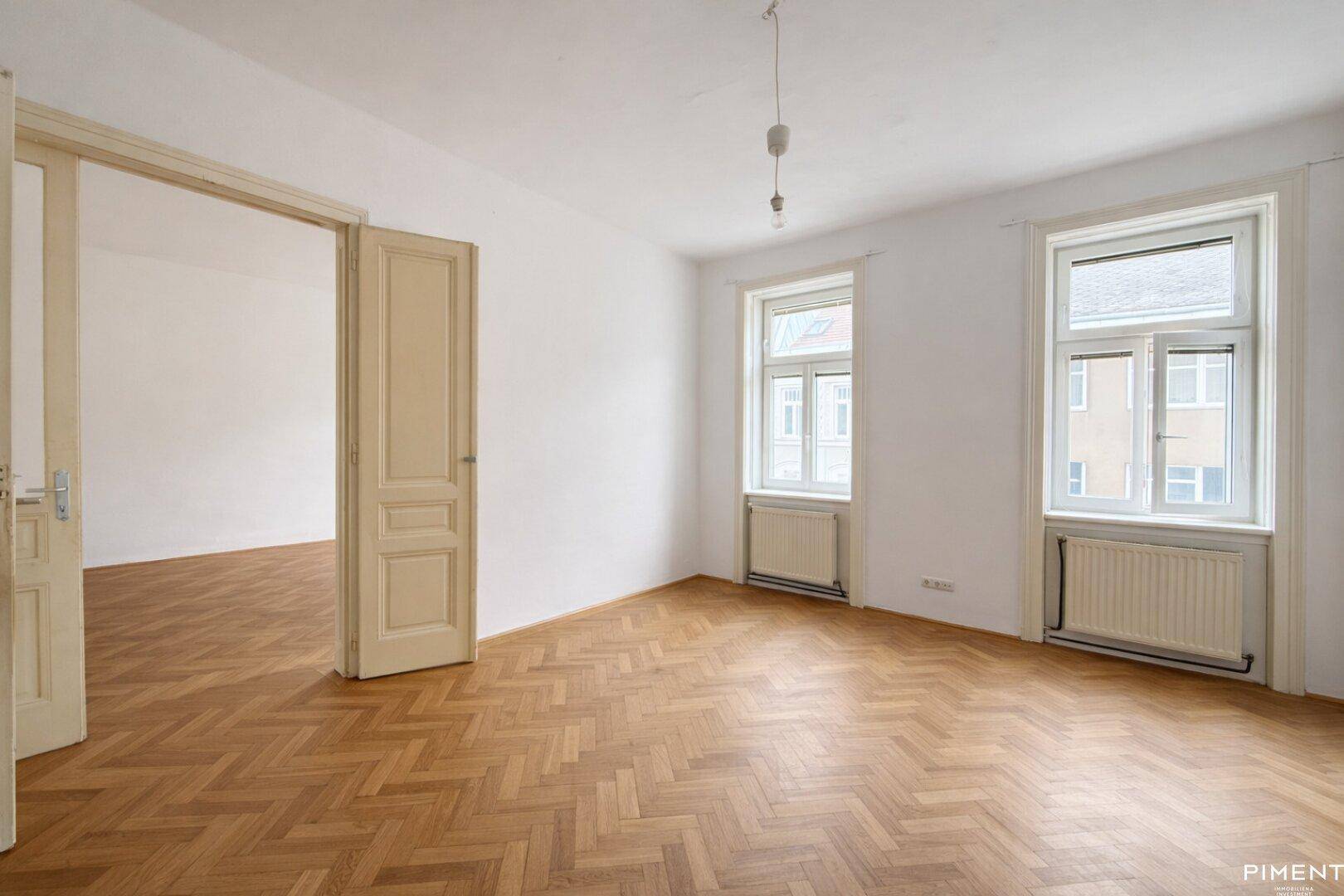 Leerer Raum mit Fischgrätparkett, hohen Decken und zwei großen Fenstern.