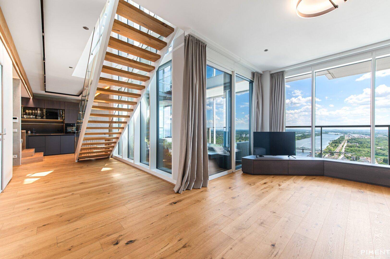 Helles Wohnzimmer mit moderner Holztreppe, großen Fenstern und Panoramablick auf die Umgebung.