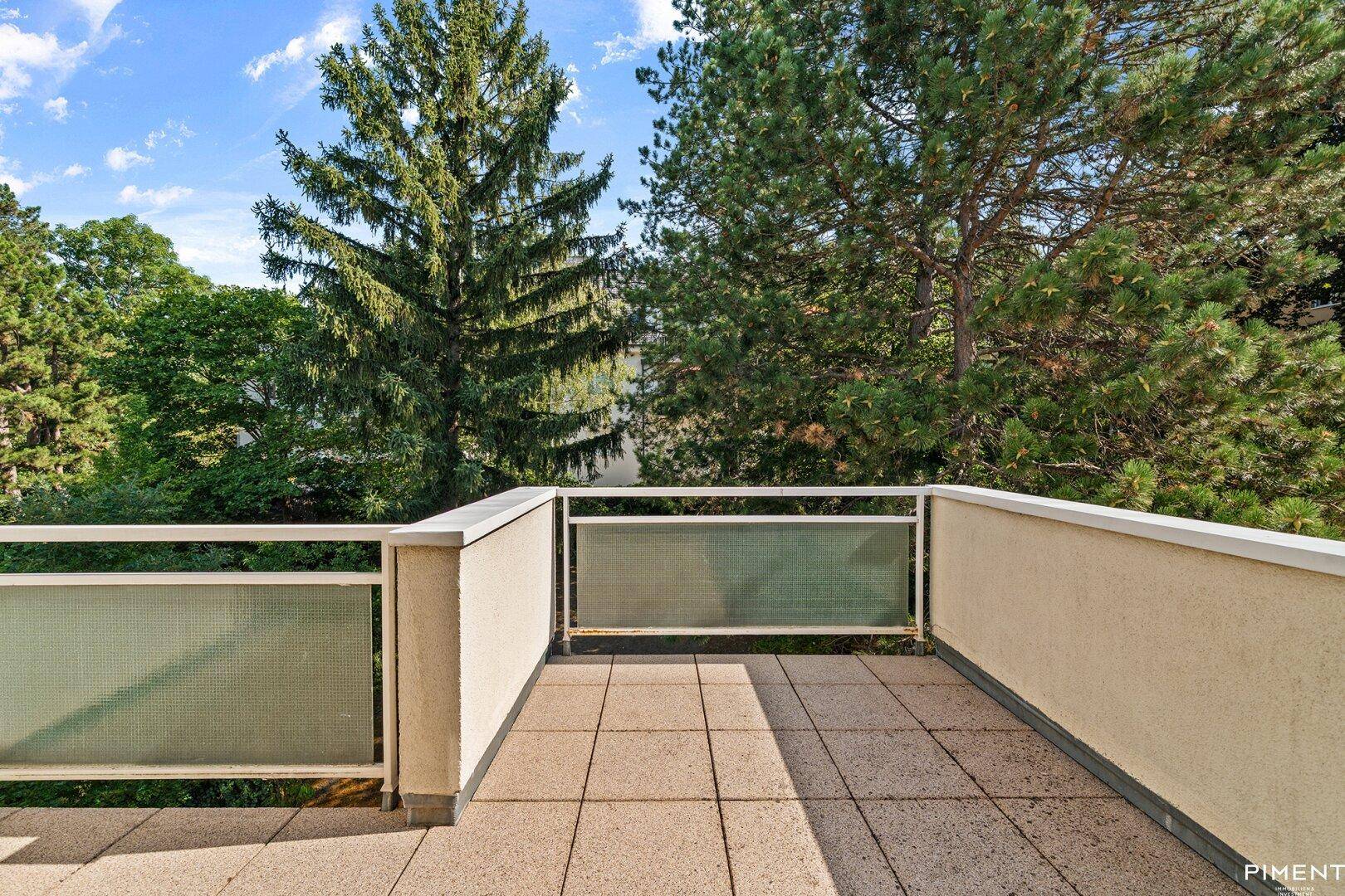 Sonniger Terrassenbereich mit Fliesenboden und Blick auf hohe Bäume und Natur.