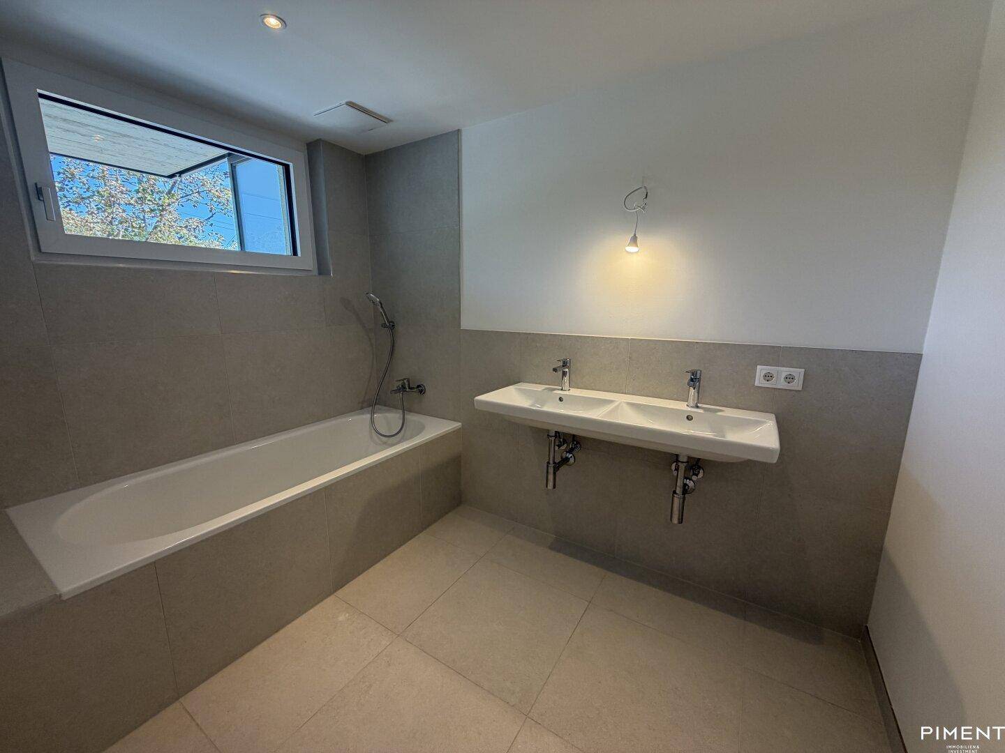 Modernes Badezimmer mit hellgrauen Fliesen, Badewanne und Doppelwaschbecken, ausgestattet mit einem Fenster.
