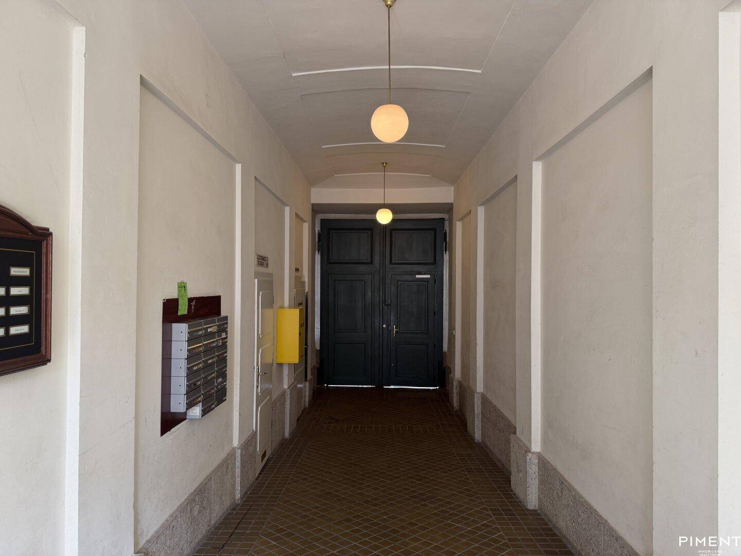 Langer, heller Flur im Gebäude mit Fliesenboden, Briefkästen und einer großen Eingangstür.