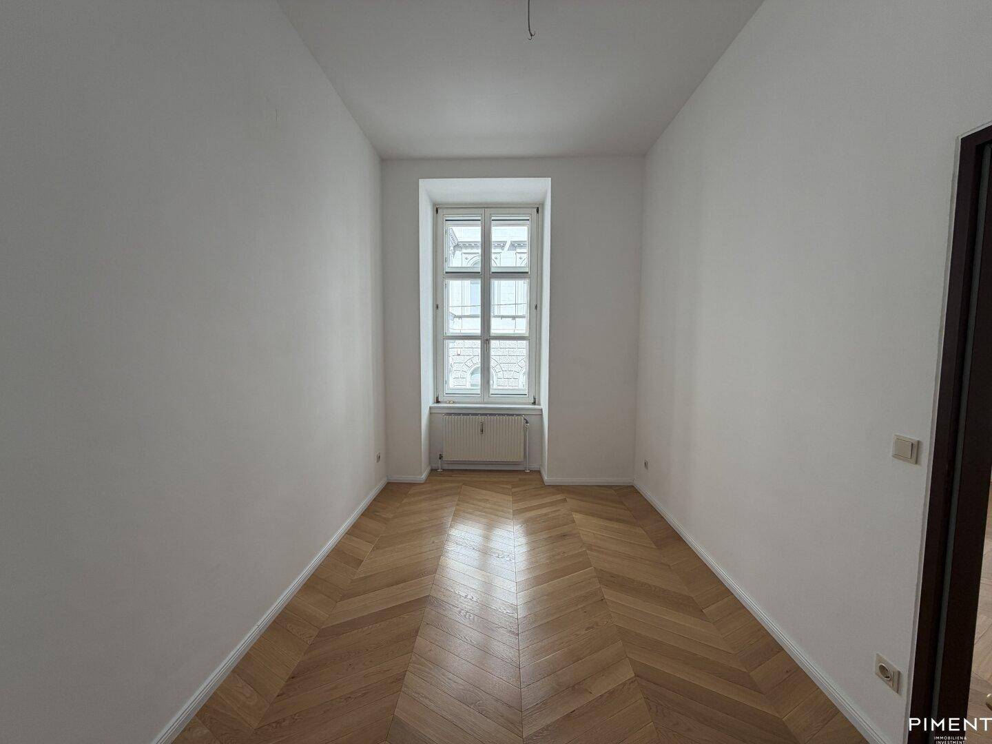 Ruhiges Zimmer mit Fischgrätparkett und einem großen Fenster, ideal als Schlaf- oder Arbeitszimmer.