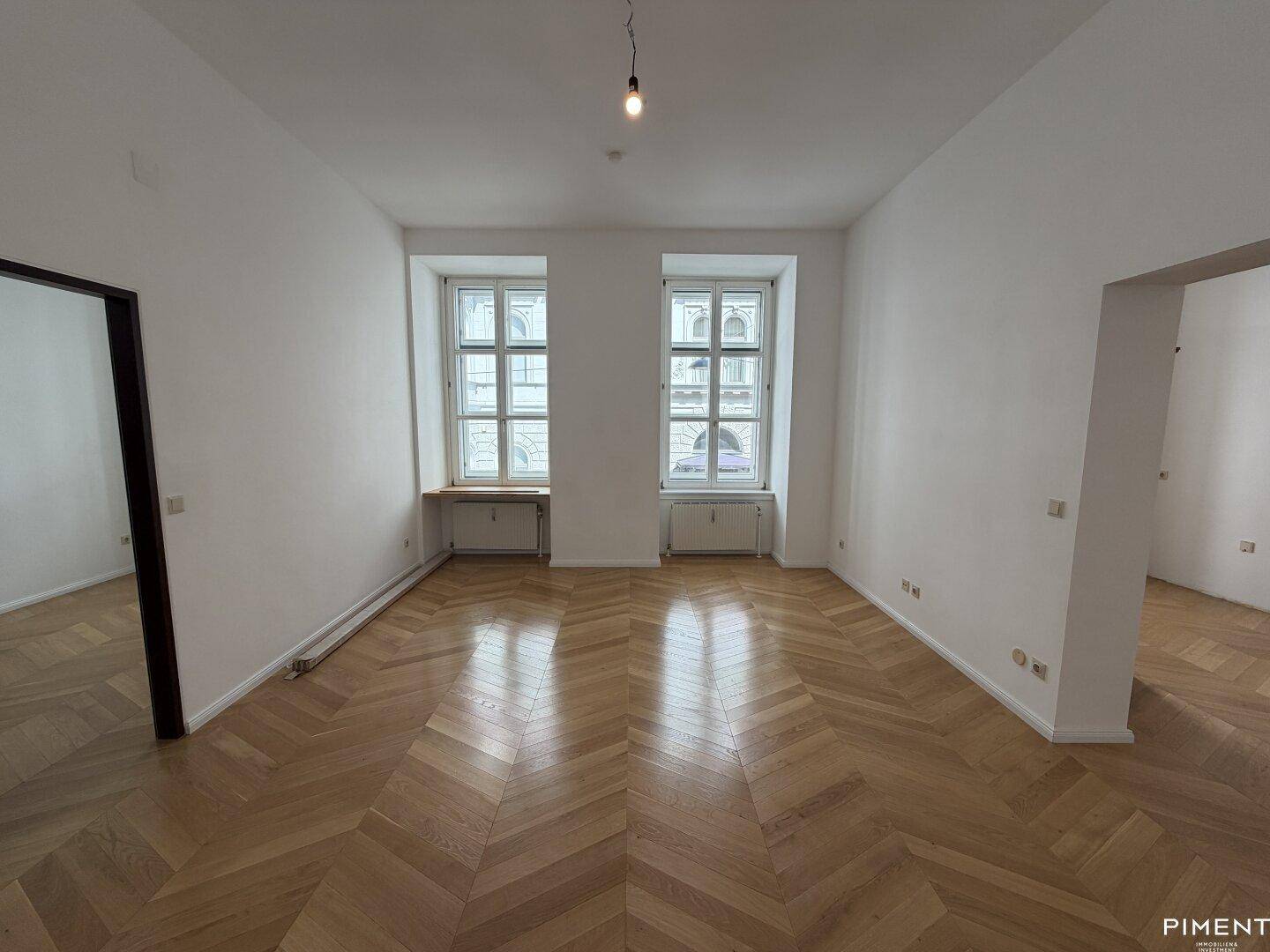 Geräumiges Zimmer mit klassischem Fischgrätparkett und zwei großen Fenstern, die den Raum erhellen.
