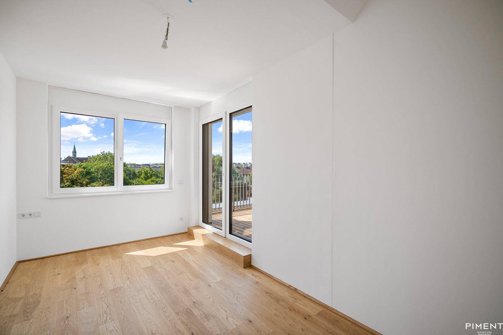 Helles Zimmer mit Holzboden, großen Fenstern und Zugang zu einem kleinen Balkon.