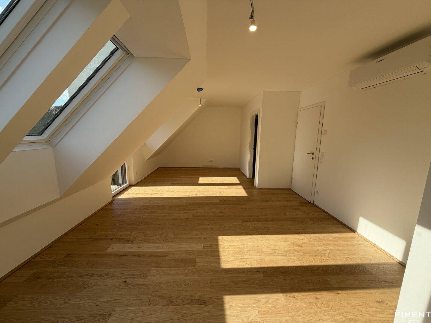 Lichtdurchfluteter Raum im Dachgeschoss mit Holzboden, Dachfenstern und Klimaanlage.