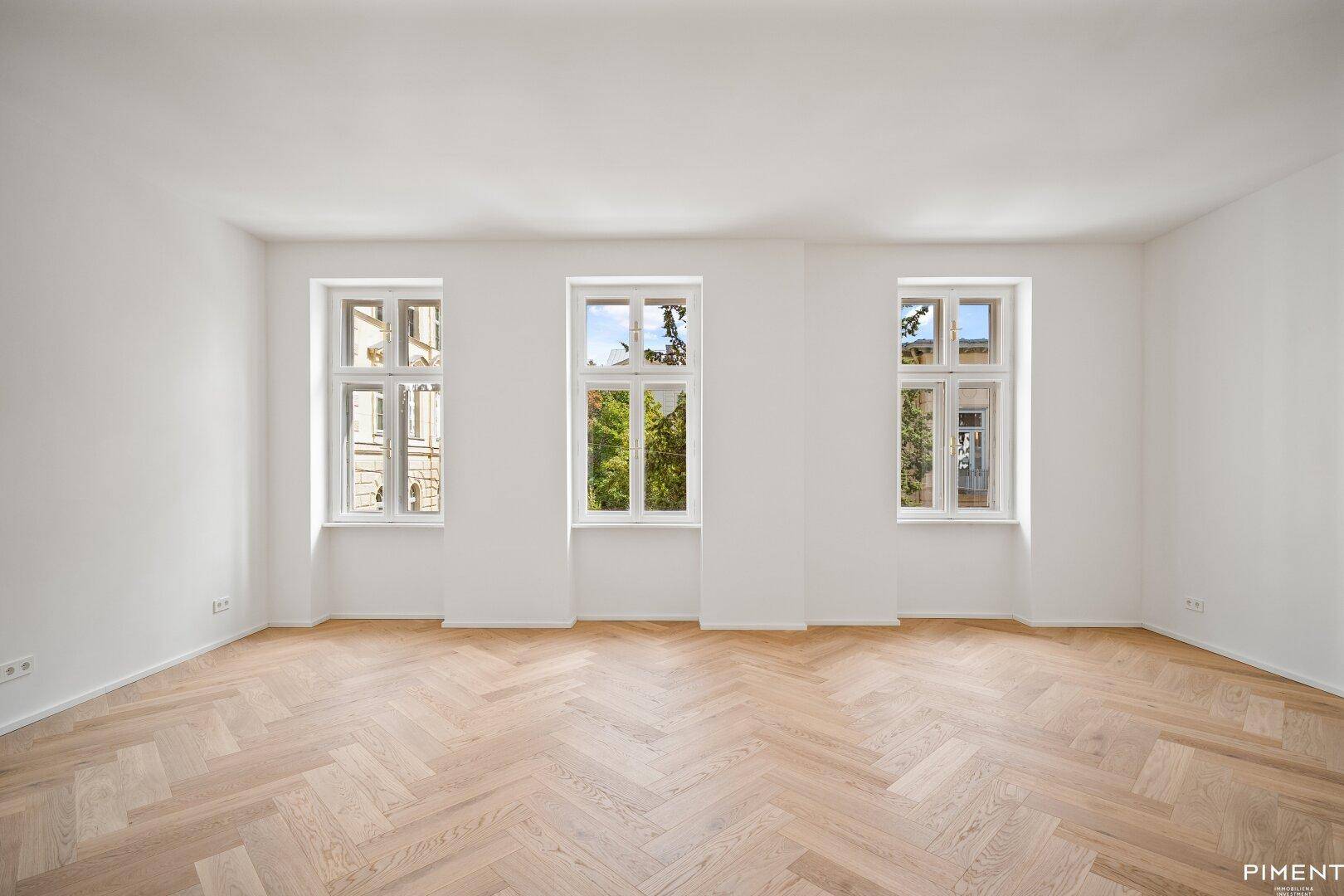 Geräumiges, unmöbliertes Zimmer mit hellem Fischgrätparkett und Blick ins Grüne.