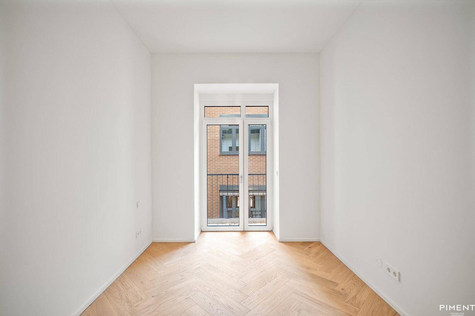 Unmöbliertes Zimmer mit Fischgrätparkett und bodentiefen Fenstern zum französischen Balkon.