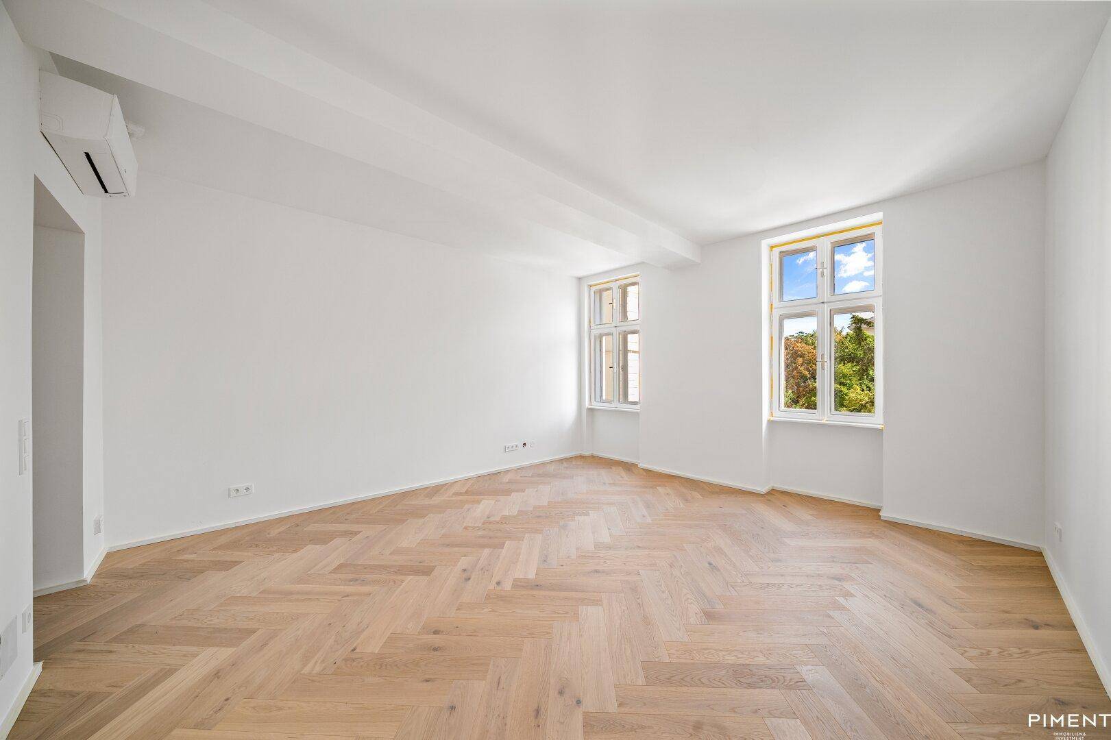 Geräumiges, helles Zimmer mit Fischgrätparkett und einer Klimaanlage an der Wand.