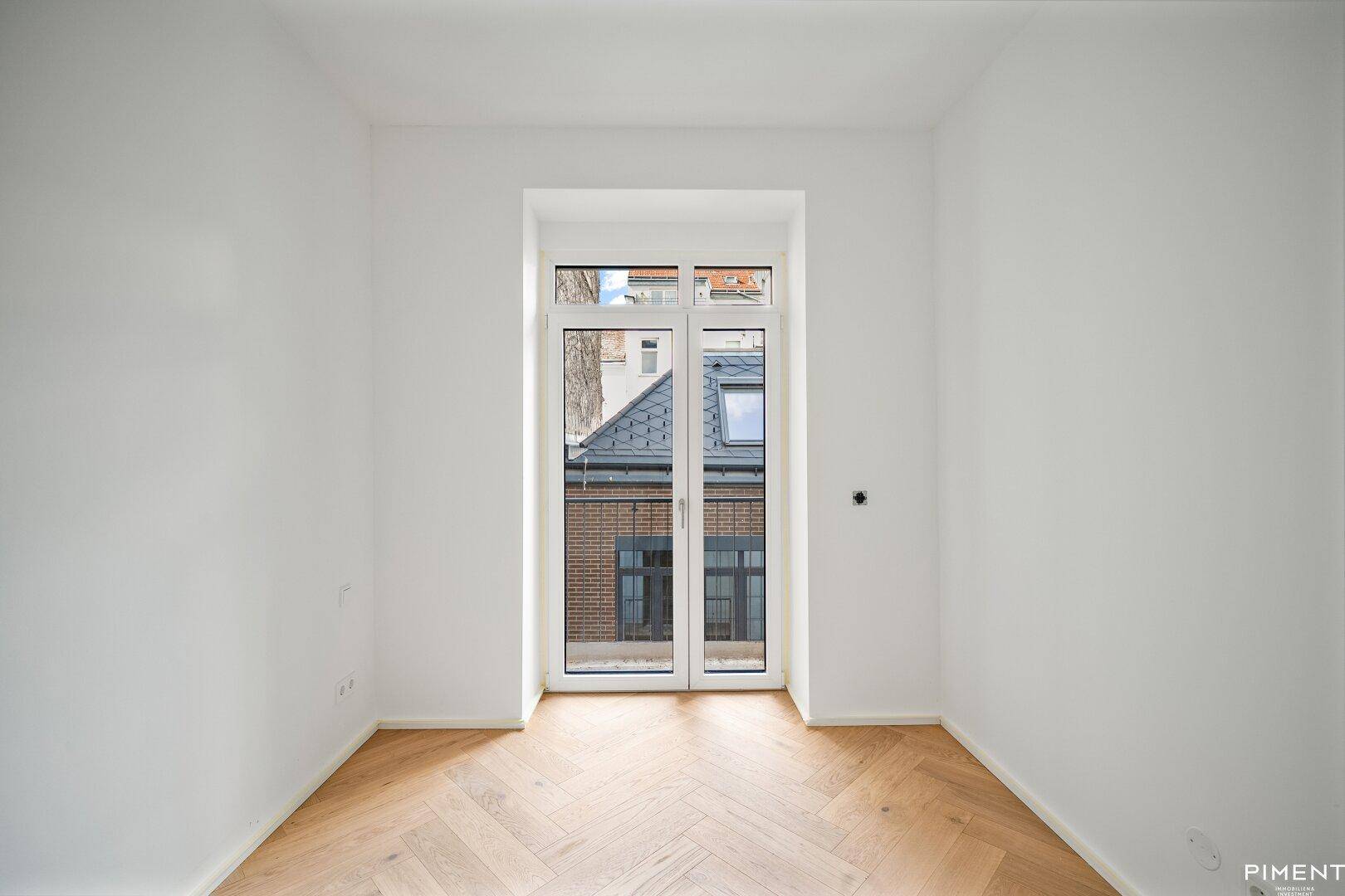 Helles Zimmer mit Fischgrätparkett und einer Fenstertür, die auf einen französischen Balkon führt.
