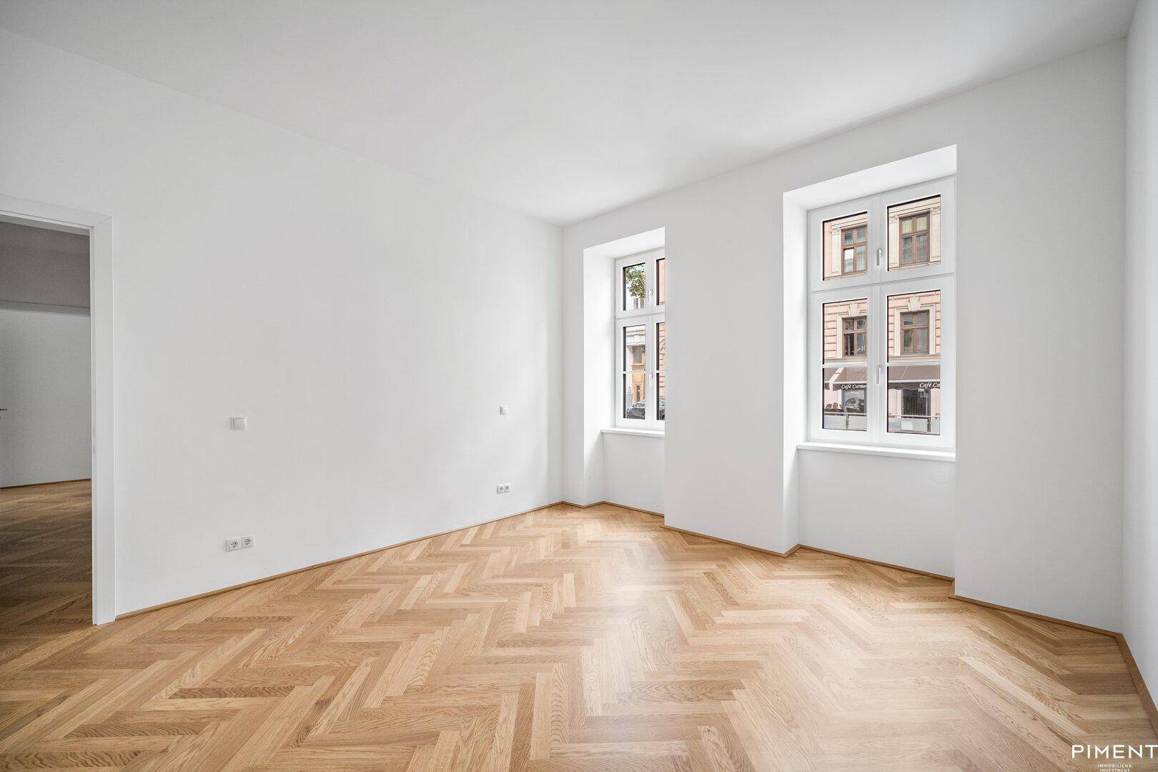 Geräumiges Zimmer mit Fischgrätparkett und zwei großen Fenstern zur Straße.
