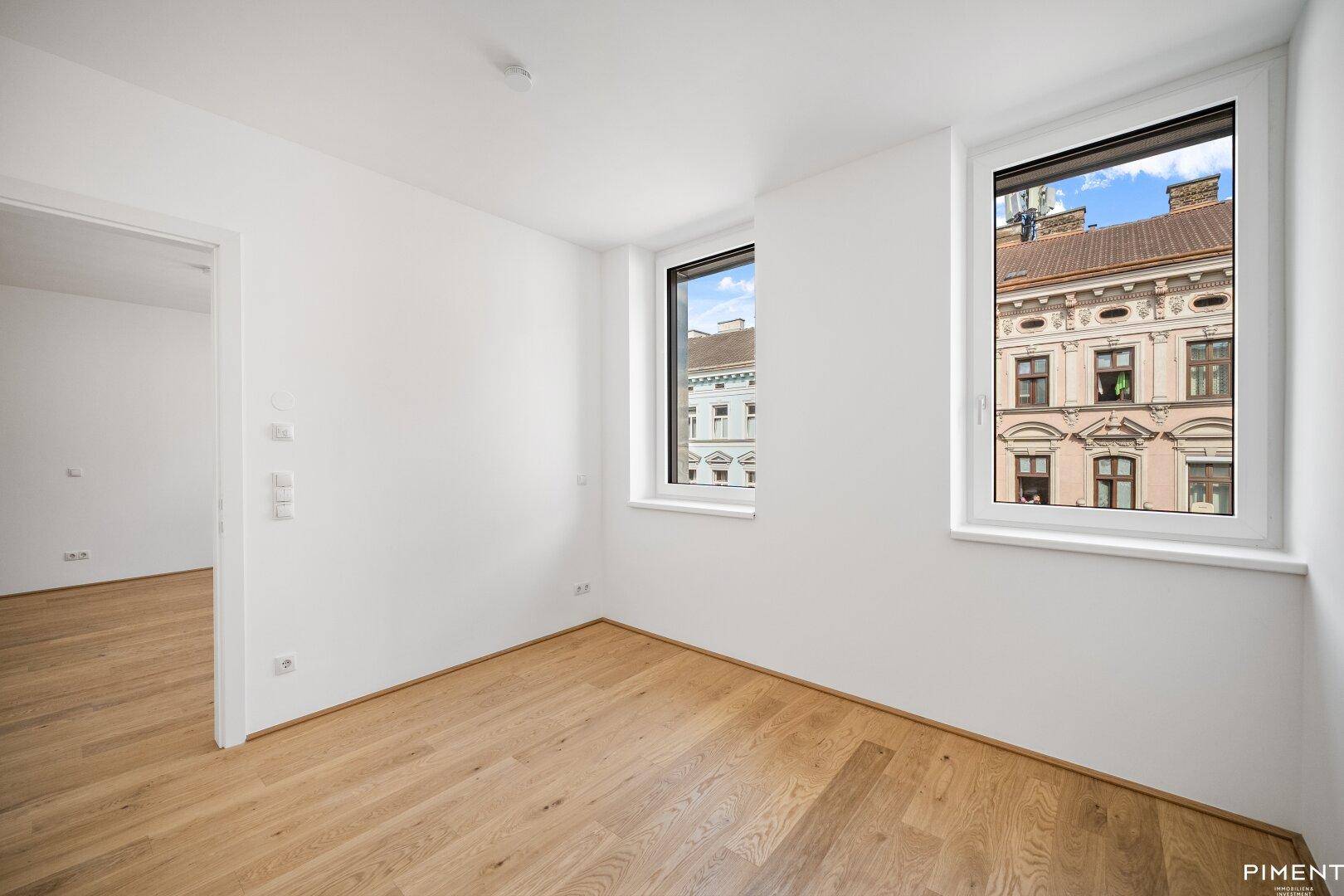 Lichtdurchflutetes, leeres Zimmer mit Holzboden und zwei Fenstern mit Stadtblick.
