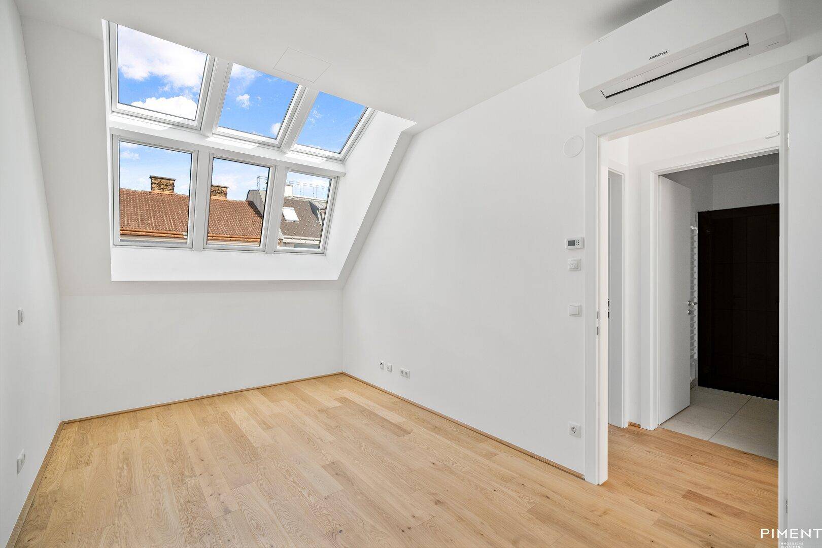 Helles Schlafzimmer mit Holzboden, Dachfenstern und Zugang zu einem weiteren Raum.
